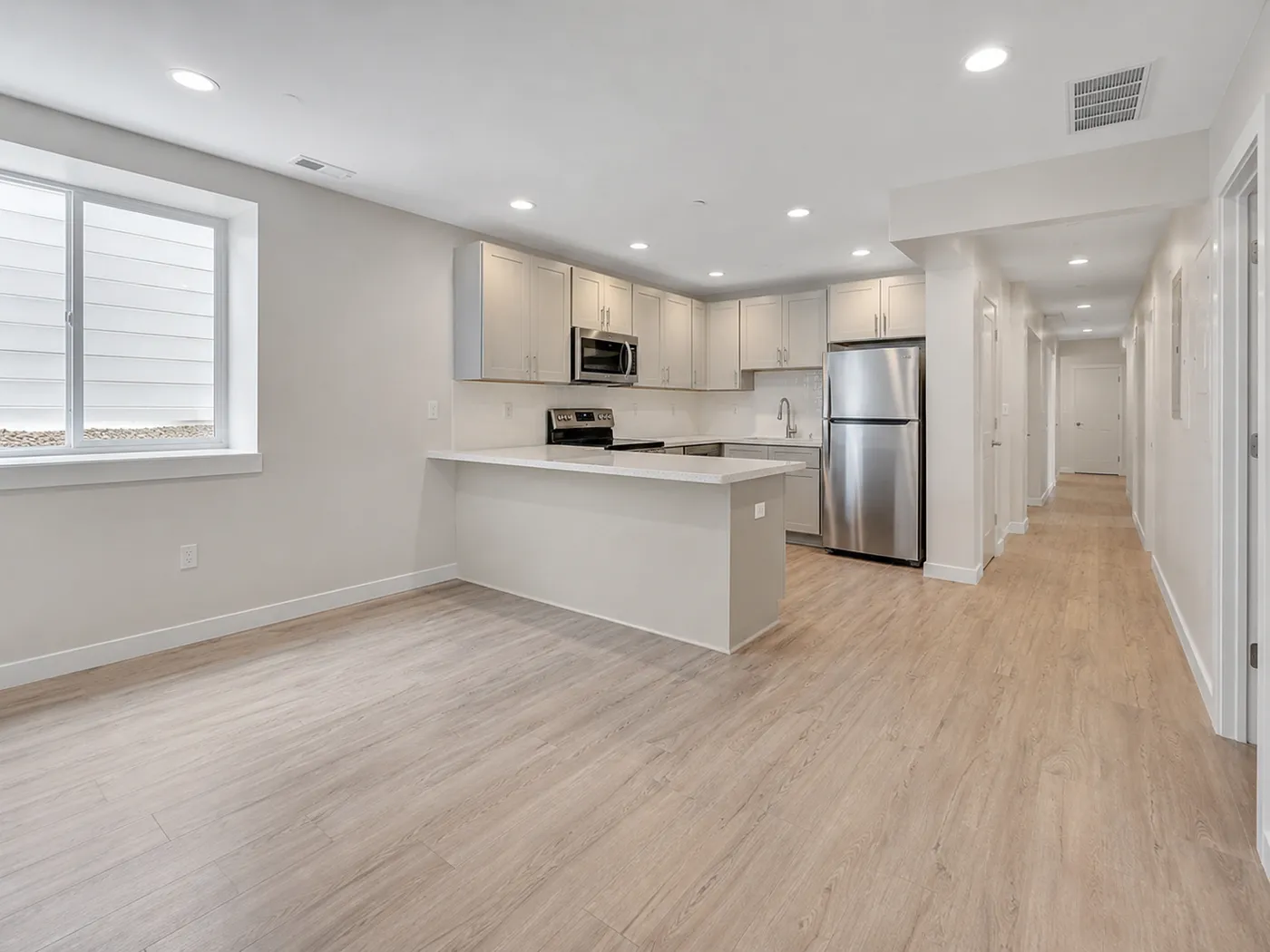 Greige basement kitchenette with peninsula seating, stainless appliances, and long daylight hallway